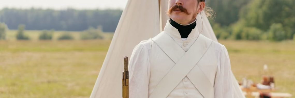 A historical reenactor in Napoleonic military uniform standing by a tent in open countryside.